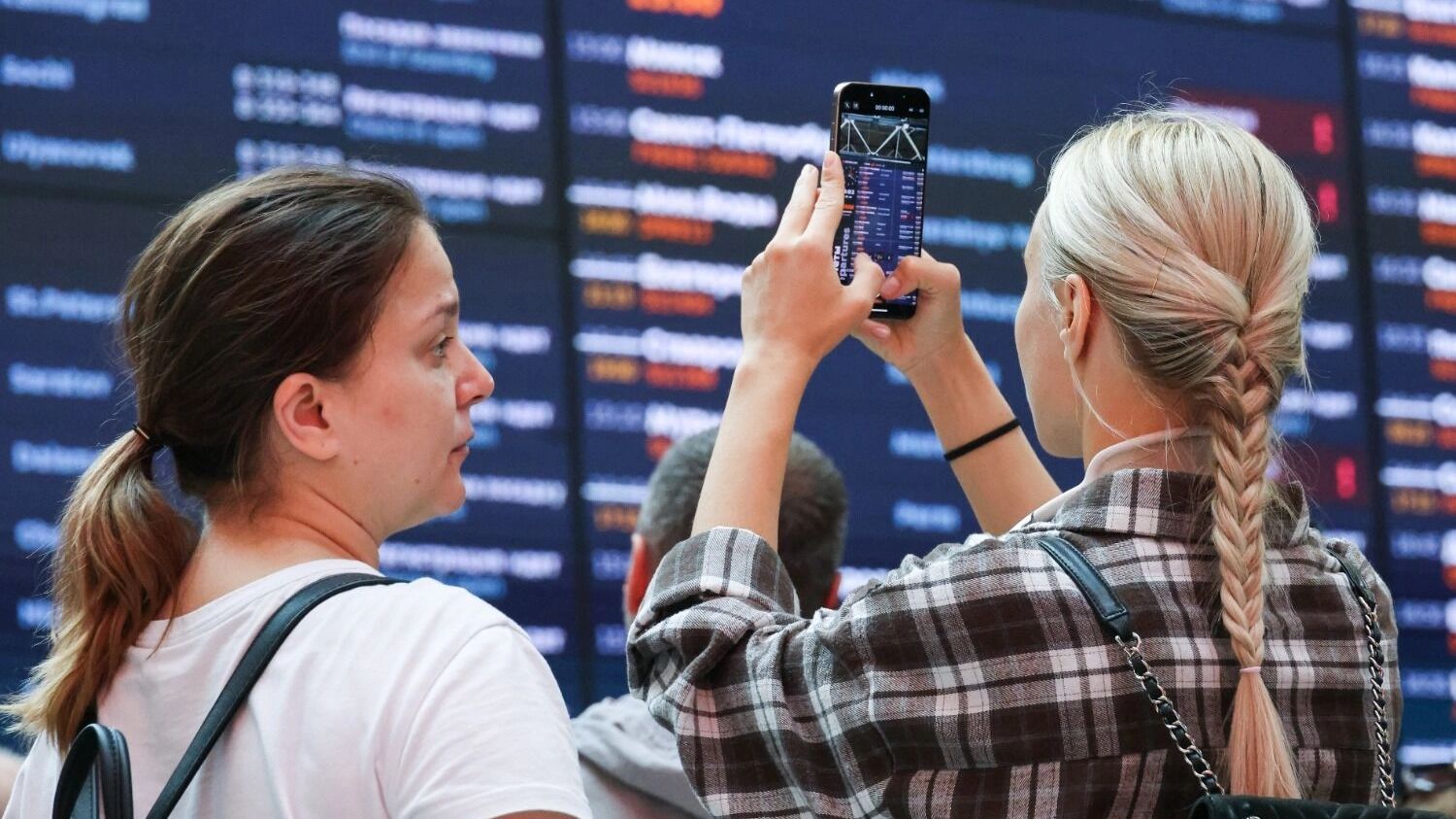 Девушки напротив табло вылетов в аэропрту снимают видео на телефон